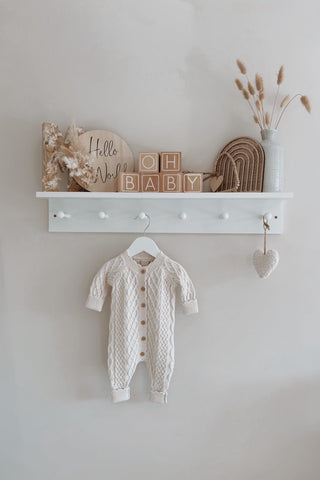 Oat cable-knit baby romper hanging in a nursery. Image also shows a shelf of blocks saying 'oh baby' 