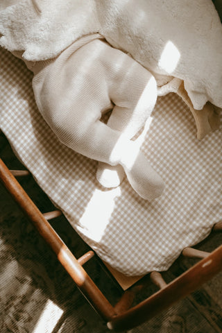 Wooden baby cot with gingham sheets. Baby feet wearing knitted baby trousers and cuddly goose. 